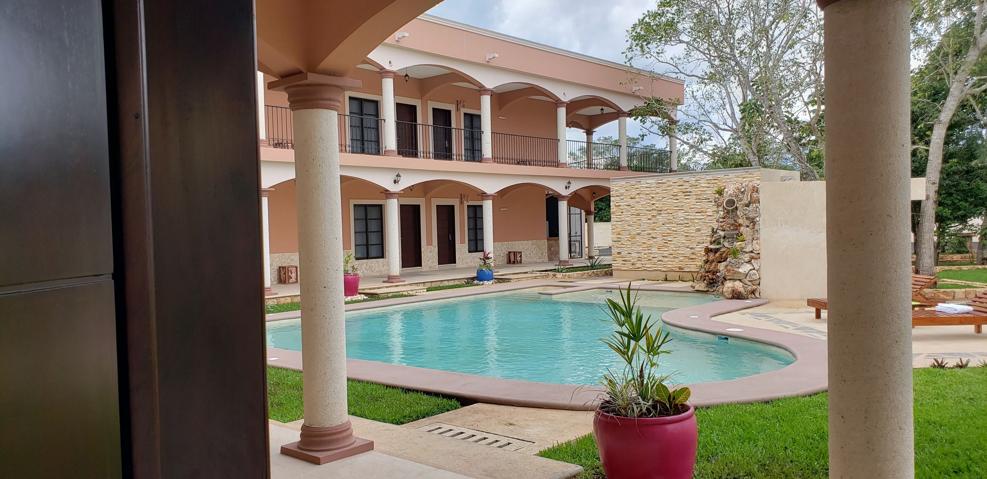 Zona de piscina con un edificio de dos pisos, columnas y un muro de piedra.