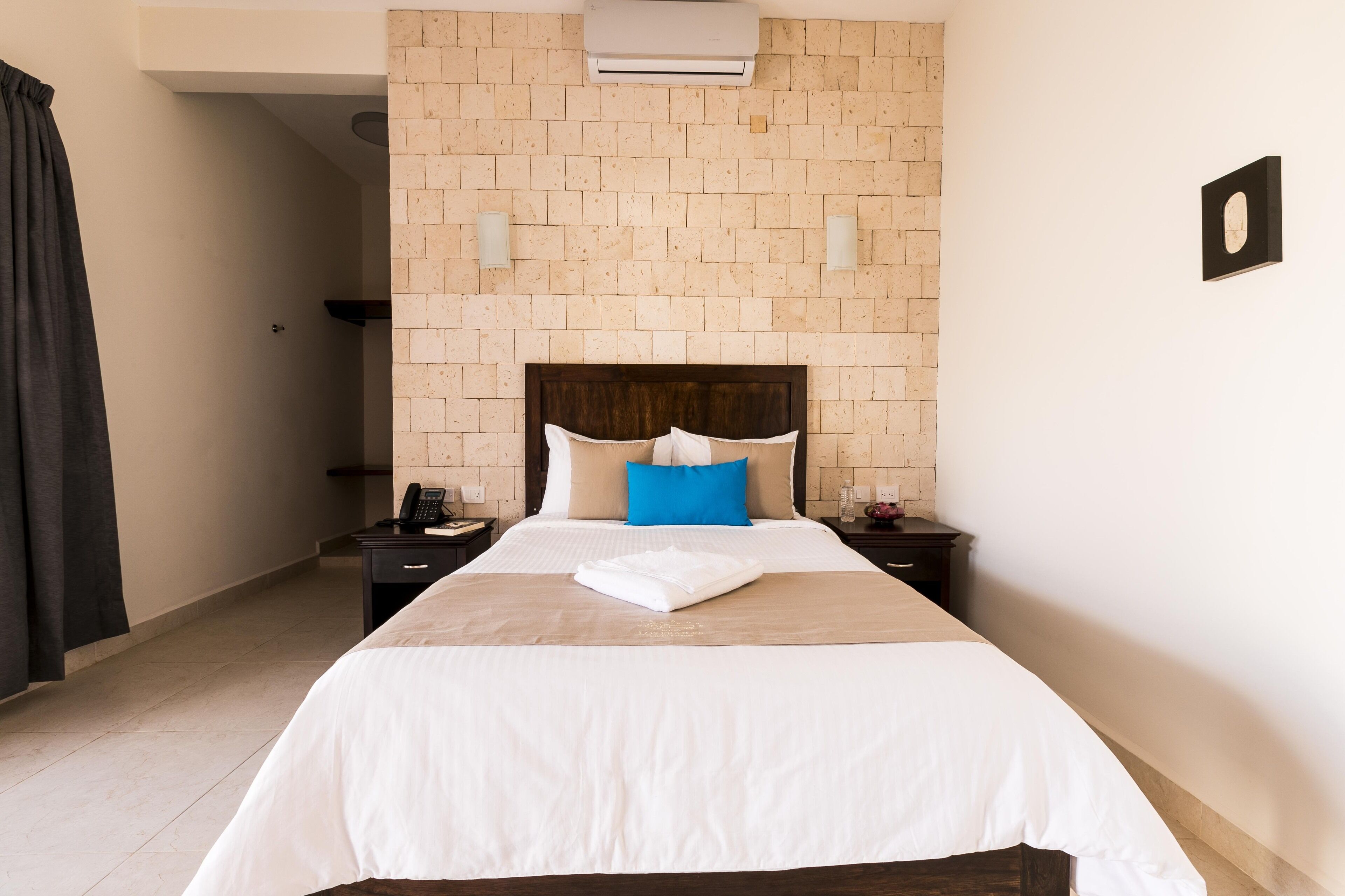 A hotel room with a brick-patterned headboard, a neatly made bed with white and blue bedding, a bedside table with a phone, and a wall-mounted air conditioner.