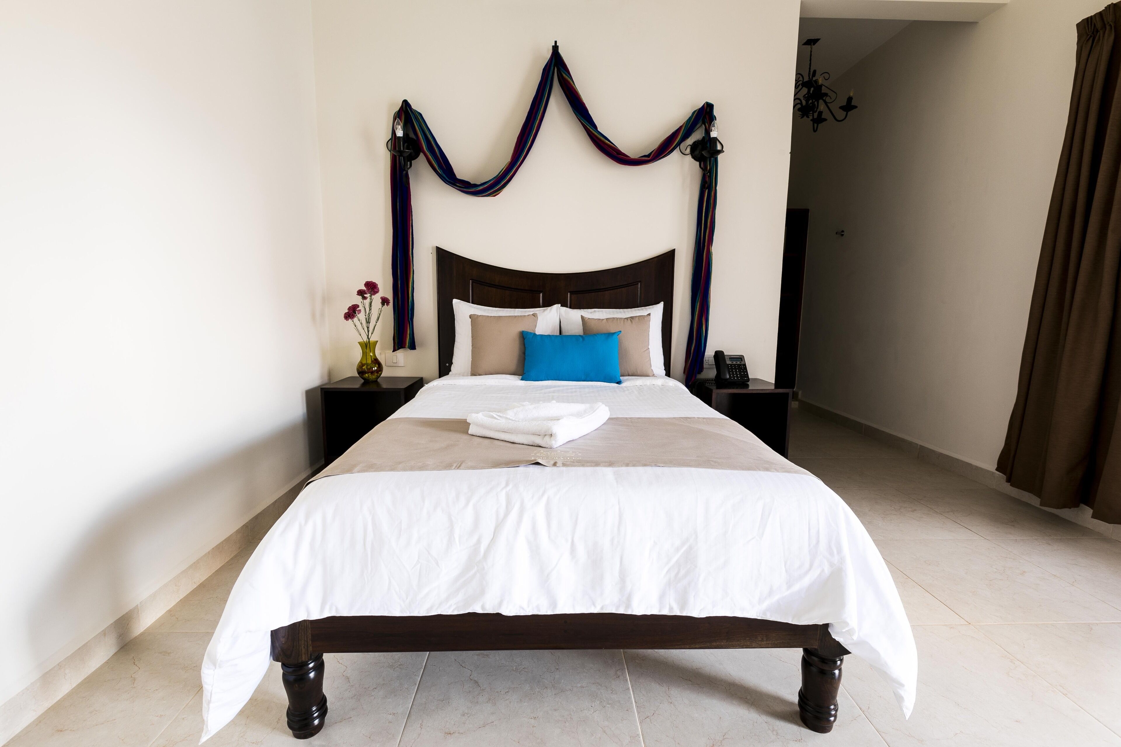 A neatly made bed with a white comforter and a blue accent pillow, a bedside table with a vase of flowers, and a dark wooden headboard.