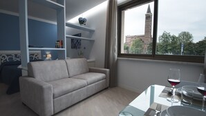 Une pièce moderne avec un canapé gris, une table à manger en verre sur laquelle se trouvent un verre à vin et une assiette, et une vue sur le clocher d’une église, visible par la fenêtre.