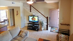 A living room with a sofa, television, ceiling fan, staircase, and a door.