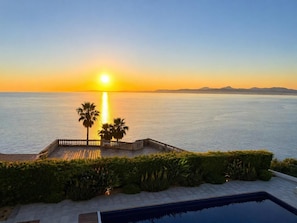 A poolside view with a sunset over the sea, palm trees, and a clear sky.