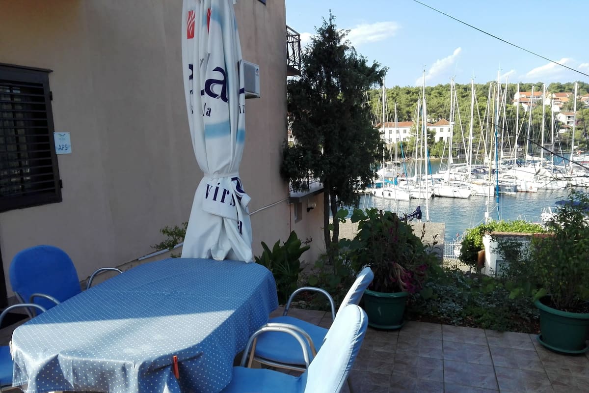 Eine Terrasse mit Tisch und Stühlen, Blick auf einen Jachthafen mit Booten und ein Gebäude mit einer Schilder.'
