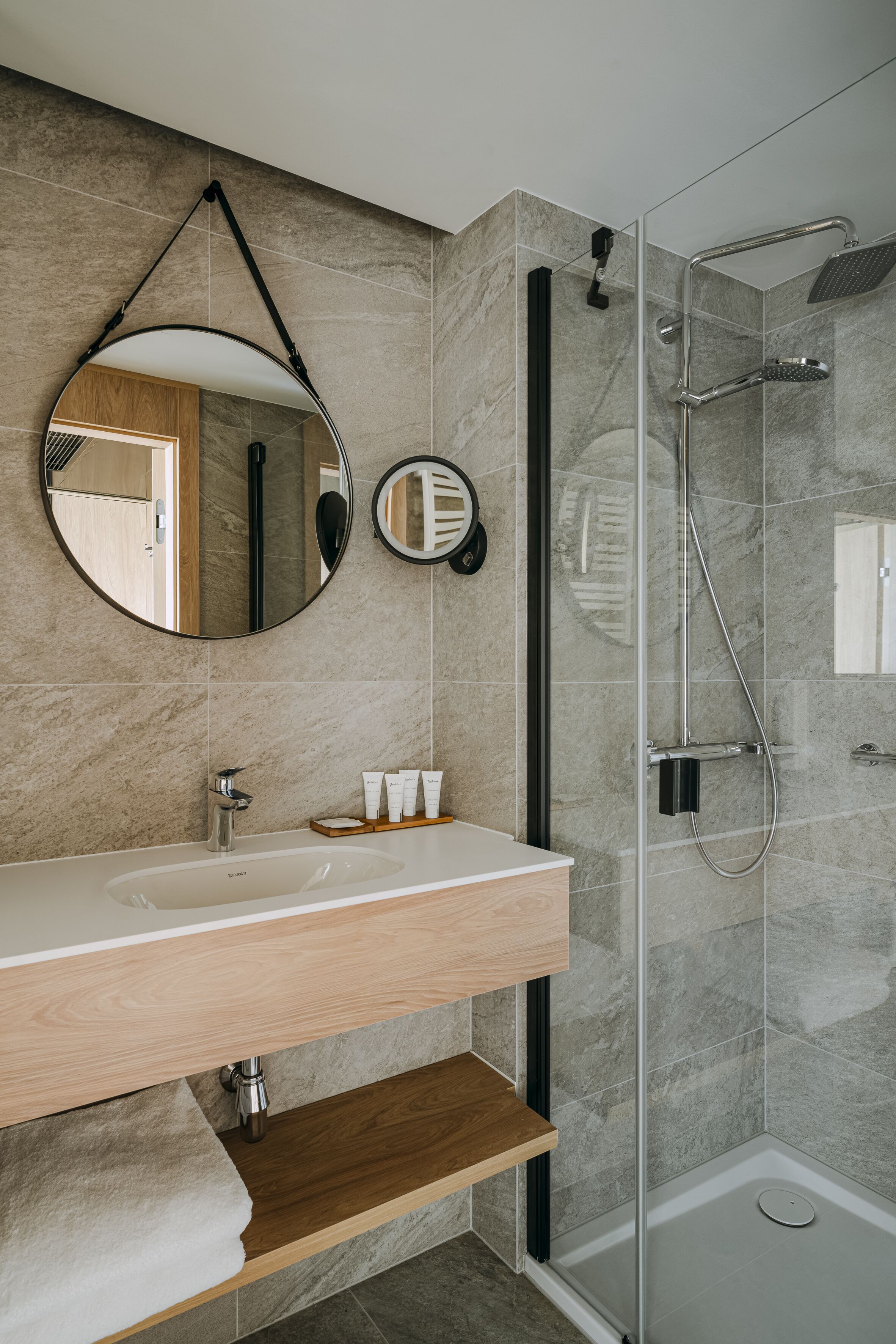 Un baño moderno con pared de mármol, un espejo redondo, ducha con cerramiento de vidrio y tocador de madera.