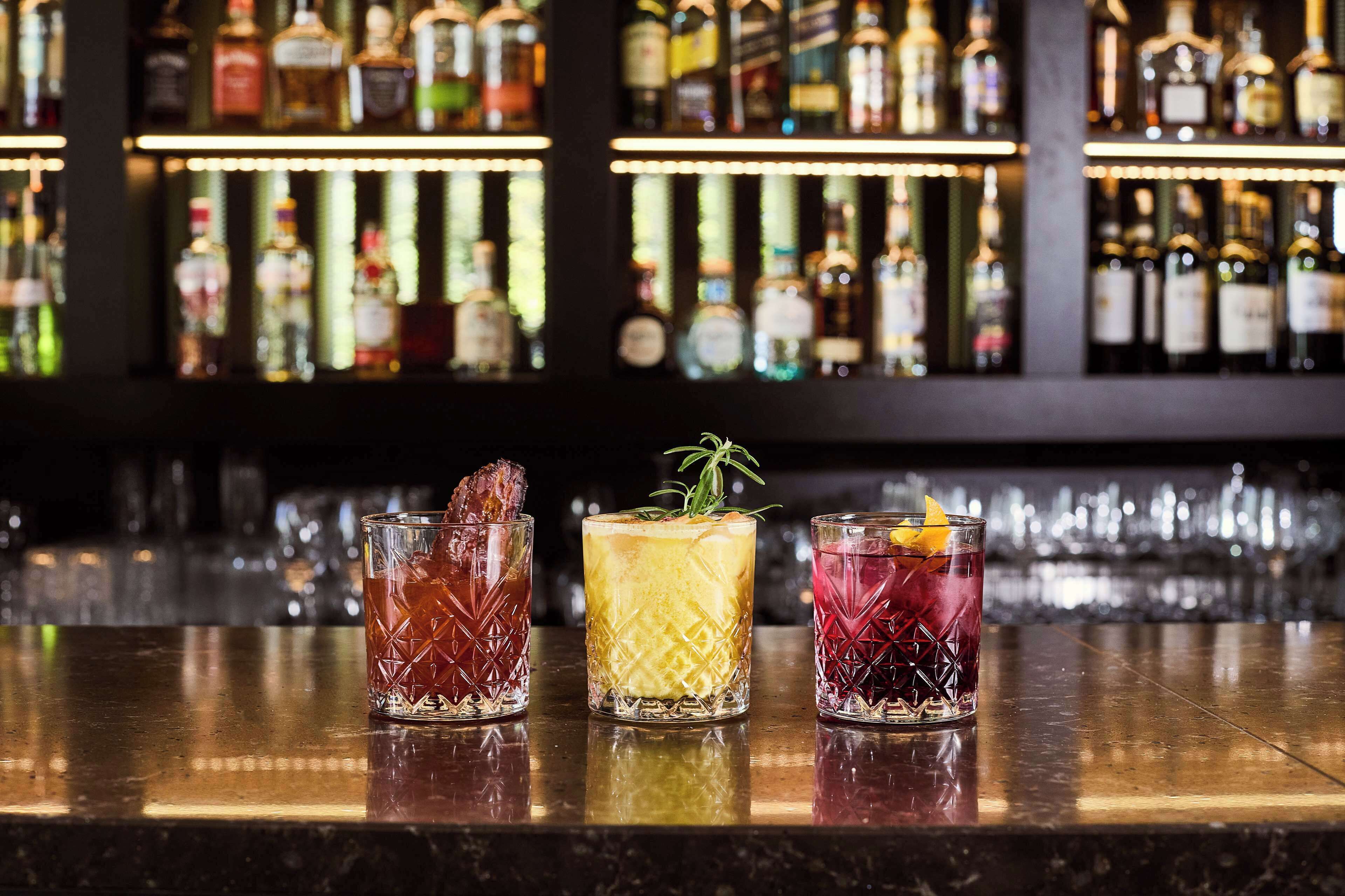 Three glasses of cocktails on a bar counter with a background of shelves stocked with bottles.
