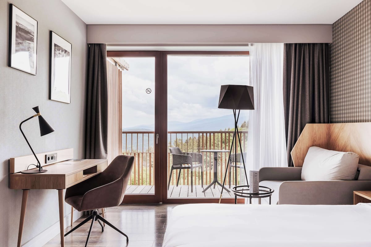 A hotel room with a bed, a desk, a chair, a sofa, a balcony with a view, and a lamp.