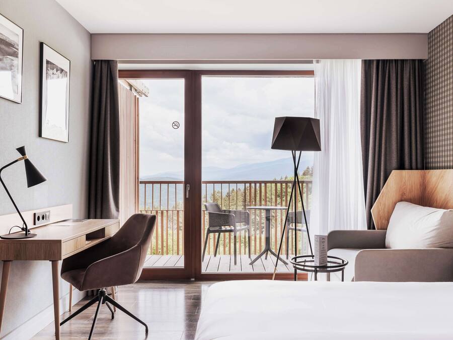 A hotel room with a bed, a desk, a chair, a sofa, a balcony with a view, and a lamp.