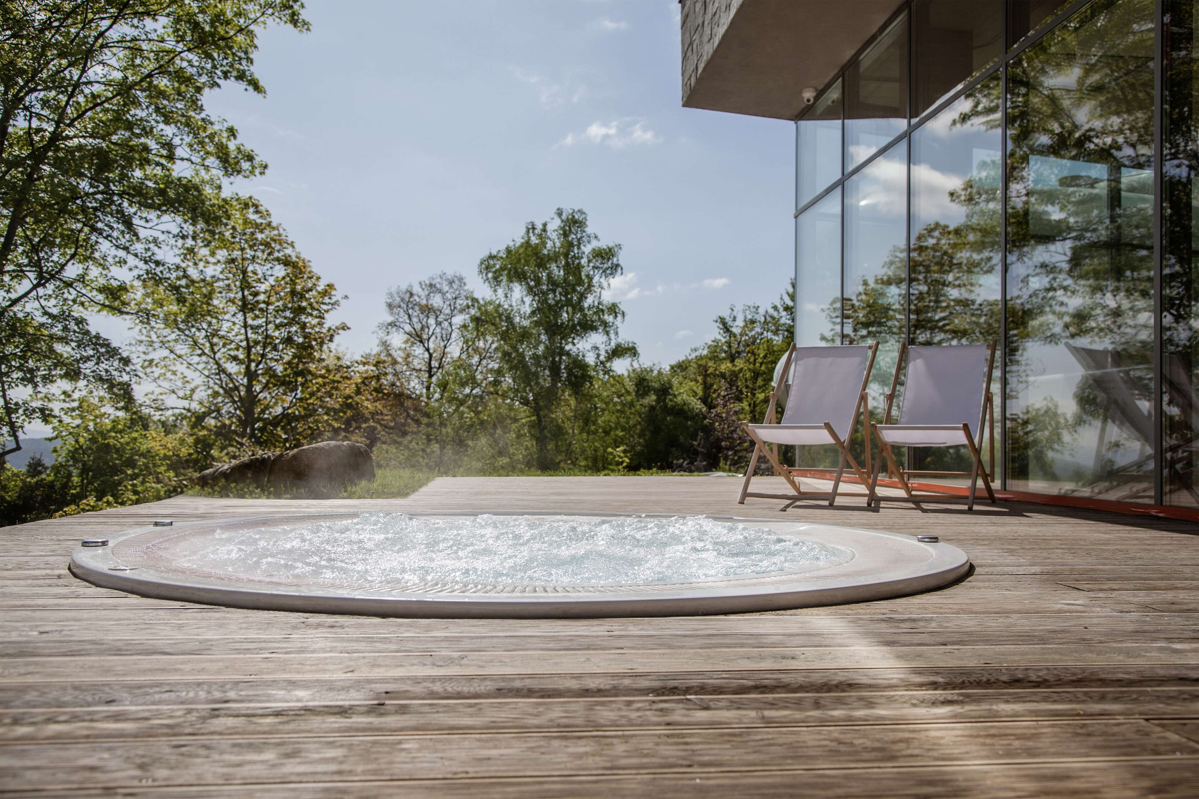 Un bain à remous sur une terrasse en bois, avec deux chaises, entouré d’arbres et d’un bâtiment en verre.