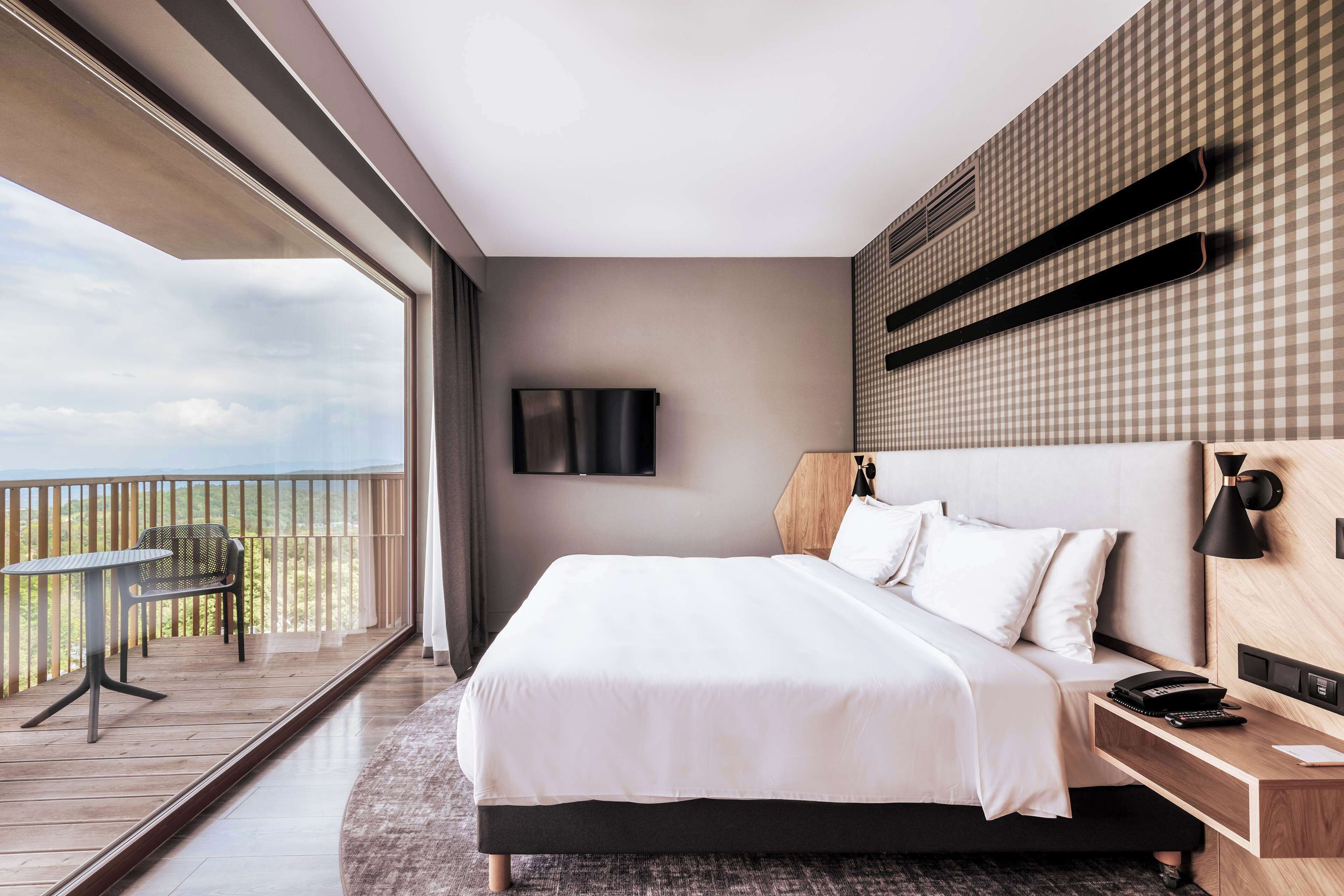 A modern hotel room with a large bed, a desk, a chair, and a balcony with a view of the sky.