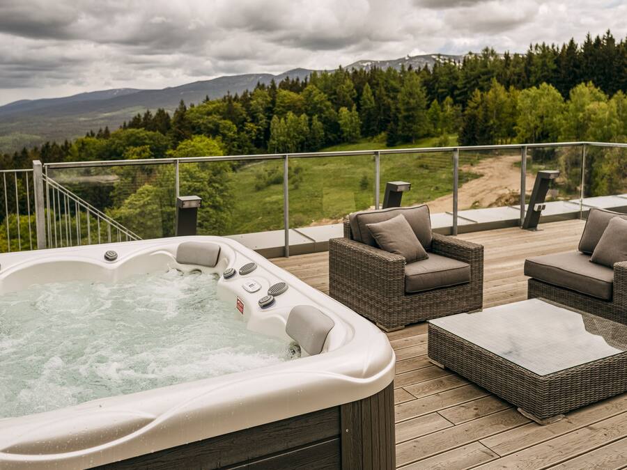 Ein Whirlpool auf einer Holzterrasse mit Korbmöbeln und Blick auf einen Wald und Berge.