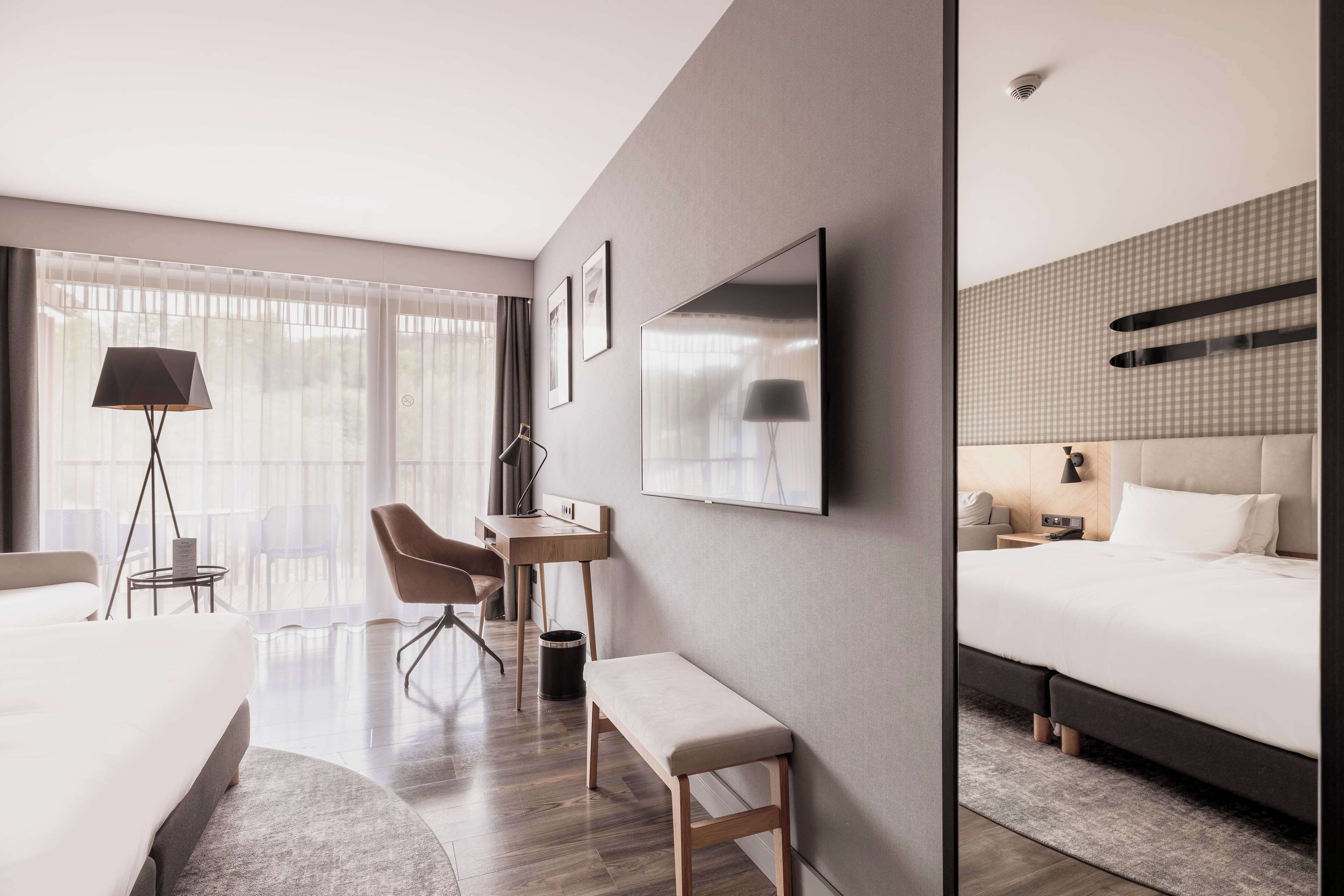 A modern hotel room with a large bed, a desk, a chair, and a TV mounted on the wall.