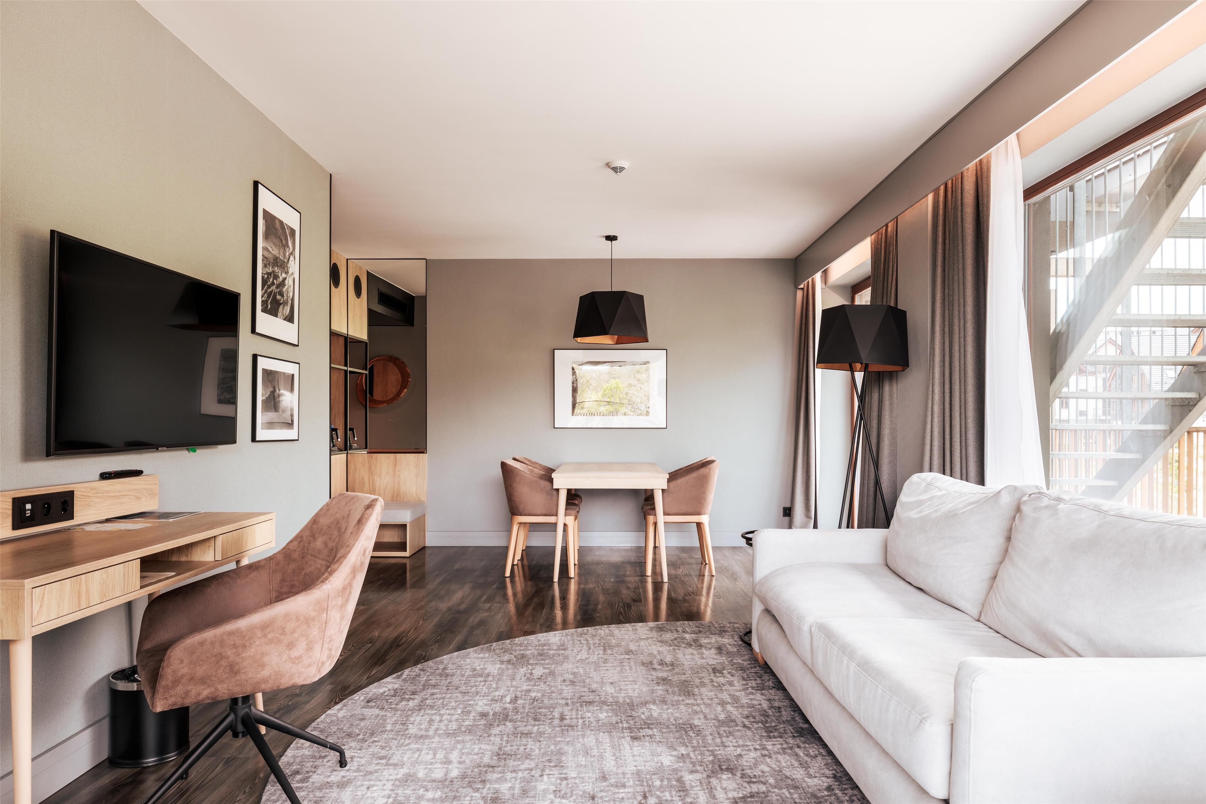 Ein modernes Hotelzimmer mit Fernseher, Schreibtisch mit Stuhl, Sofa, Esstisch mit Stühlen und einem großen Fenster mit Vorhängen.