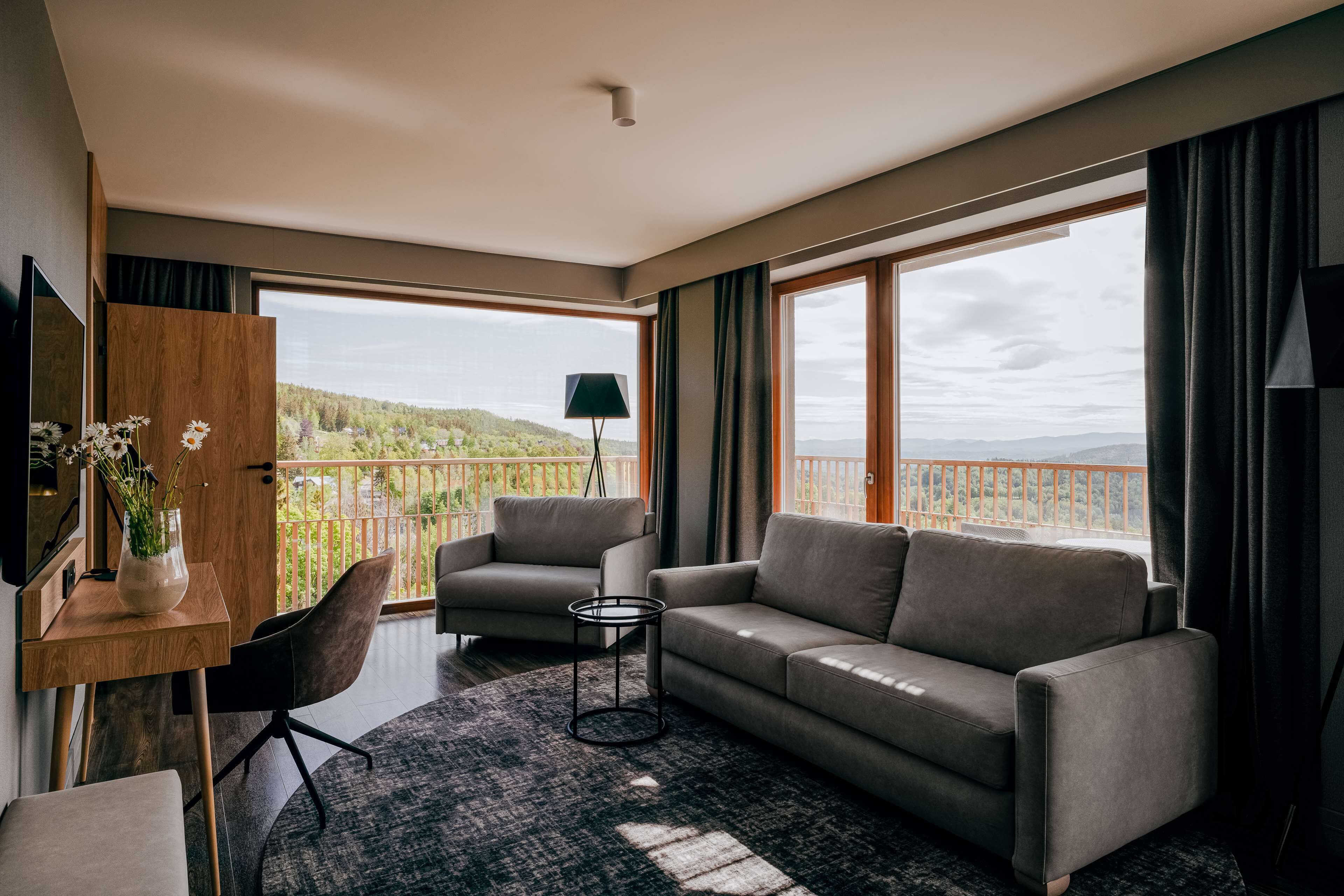 Una sala de estar moderna con un sofá, sillas, una mesita y un televisor. Amplias ventanas ofrecen vistas a zonas verdes y montañas.