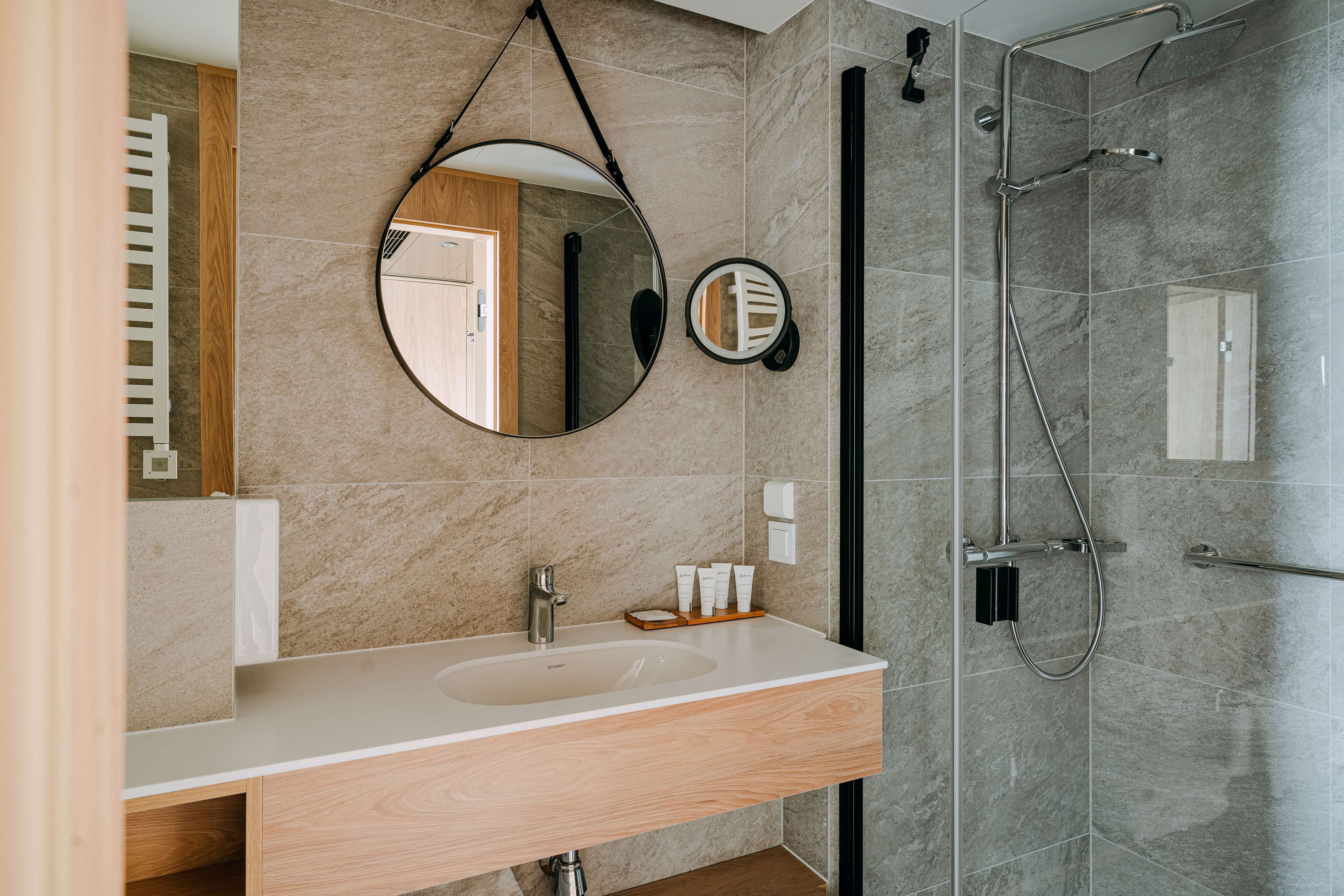 Un baño moderno con tocador de madera, espejo redondo y cabina de ducha con mampara de vidrio.
