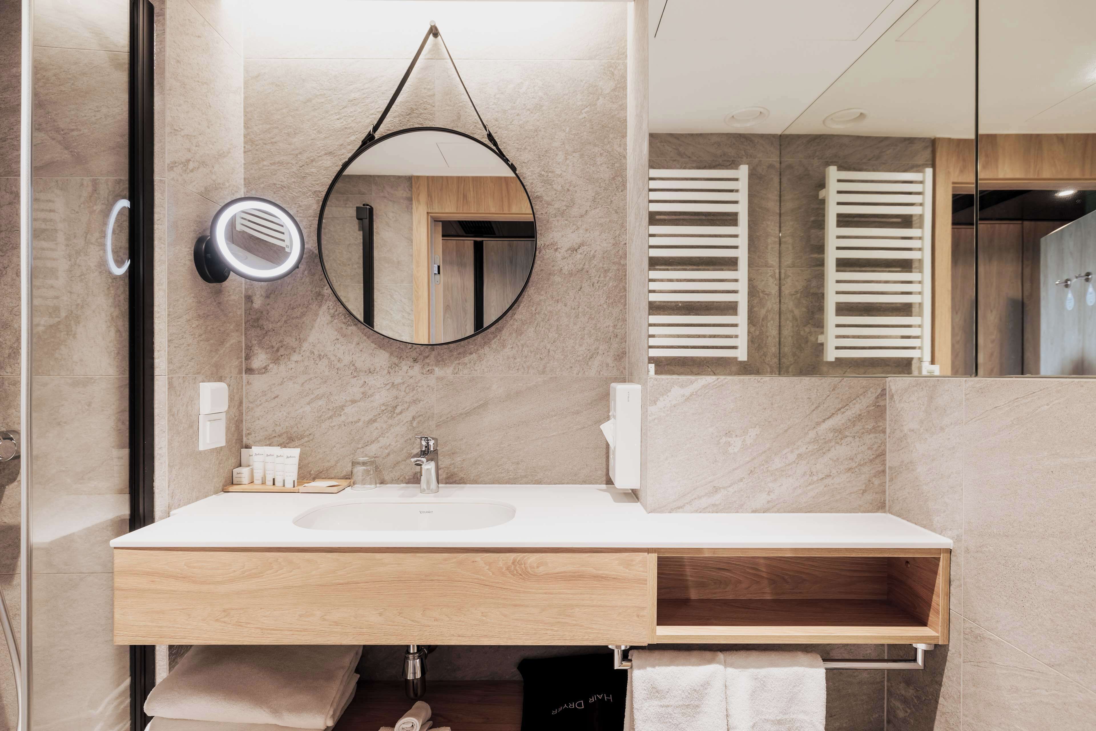 A modern bathroom with a large mirror, a round hanging mirror, a sink, and a wooden vanity.