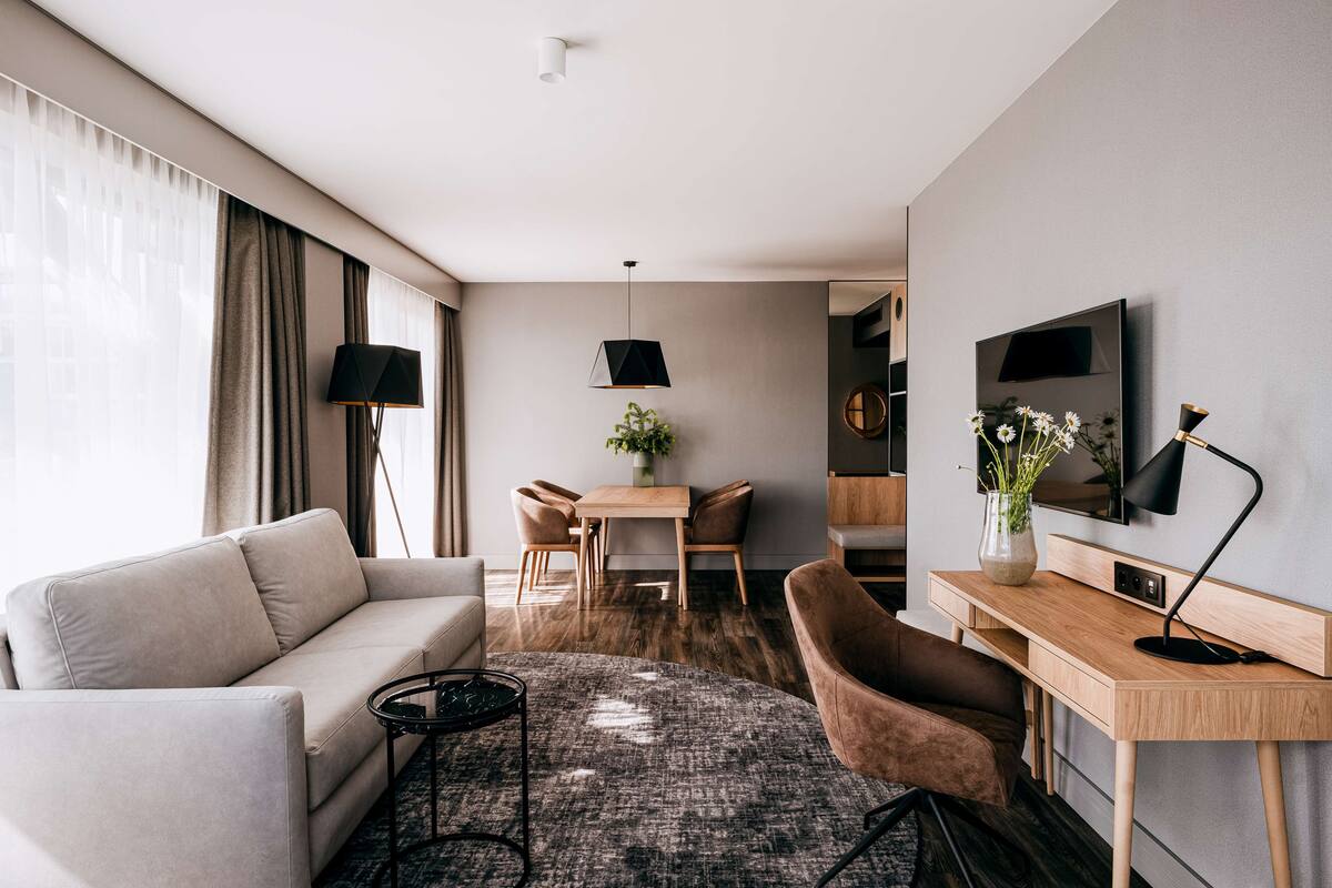 A modern hotel room with a sofa, a dining table with chairs, a desk with a computer, a TV, and a large window with curtains.