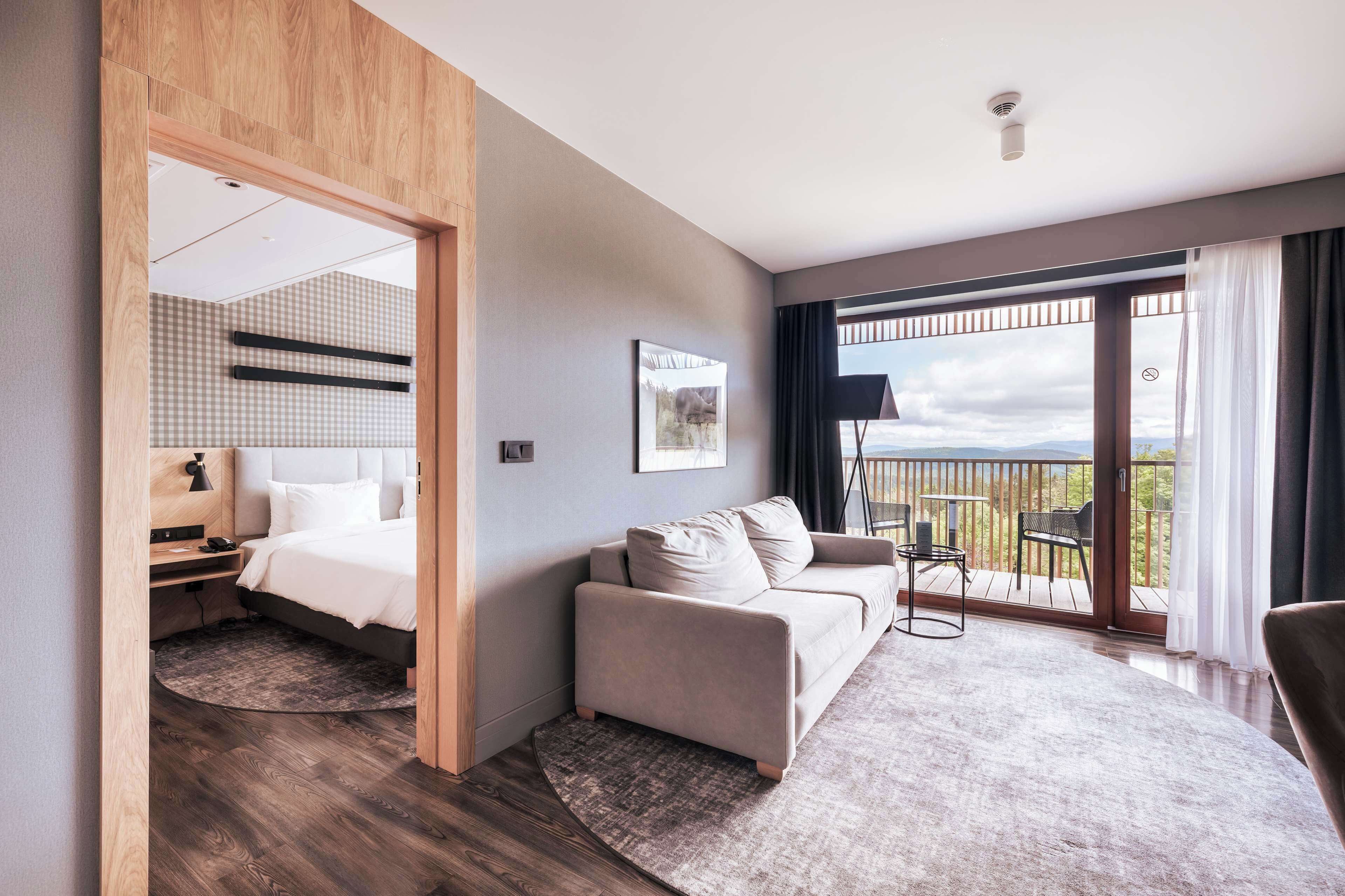 A modern hotel room with a bed, a sofa, a balcony with a view, and a desk with a lamp.
