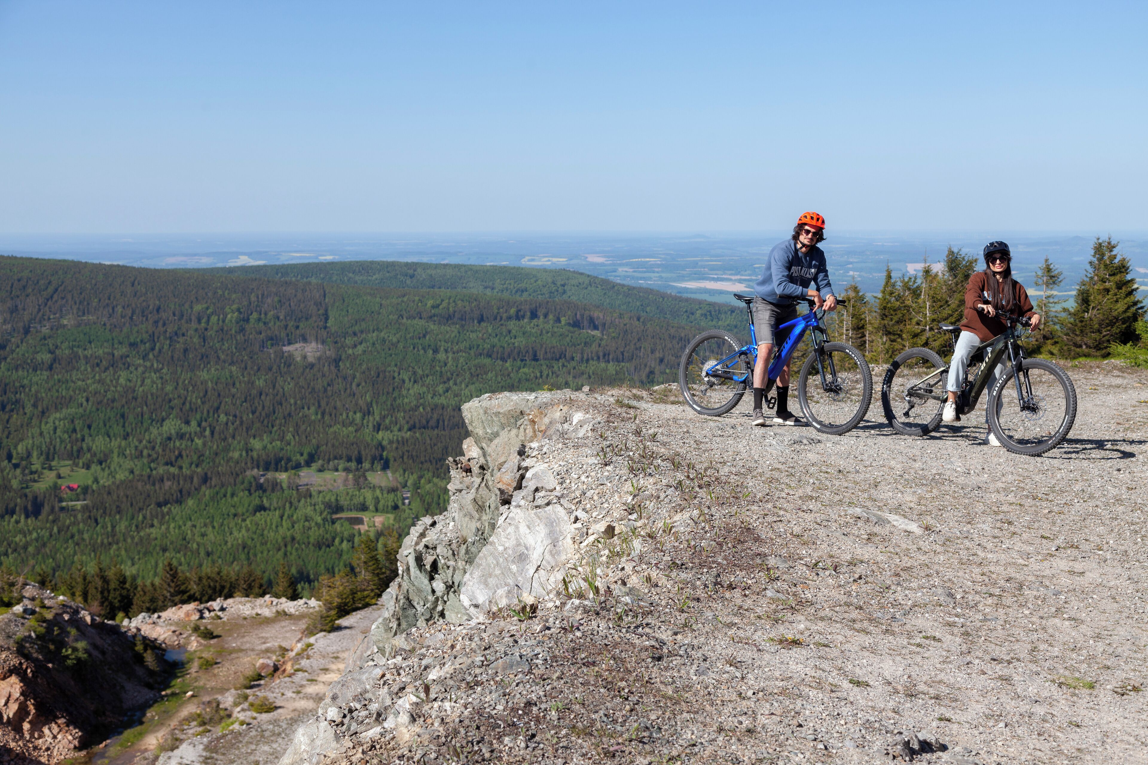 Two people on a rocky cliff with a view of a forested valley below.
