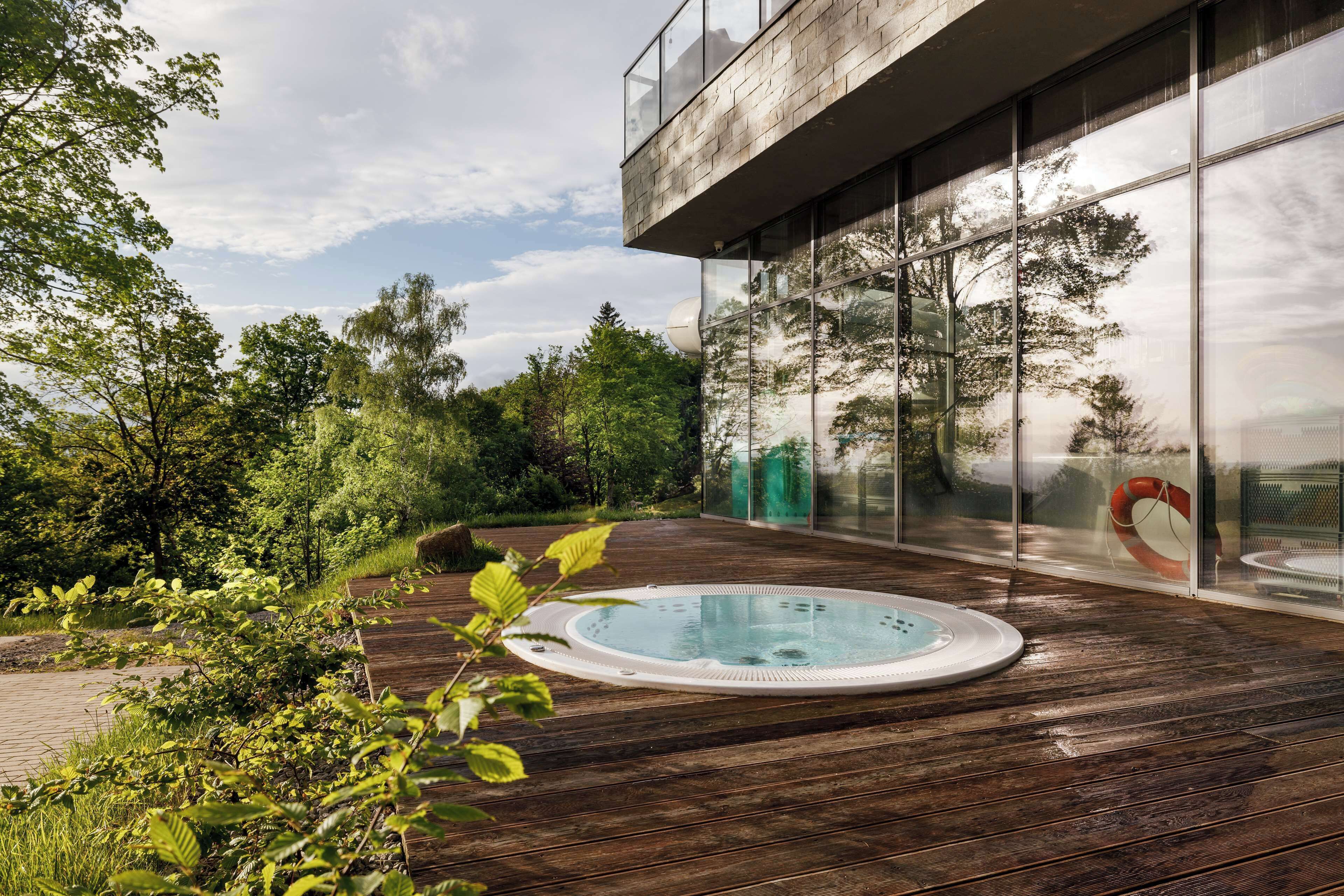 A modern outdoor pool area with a wooden deck, a hot tub, and a glass-enclosed space reflecting trees and sky.