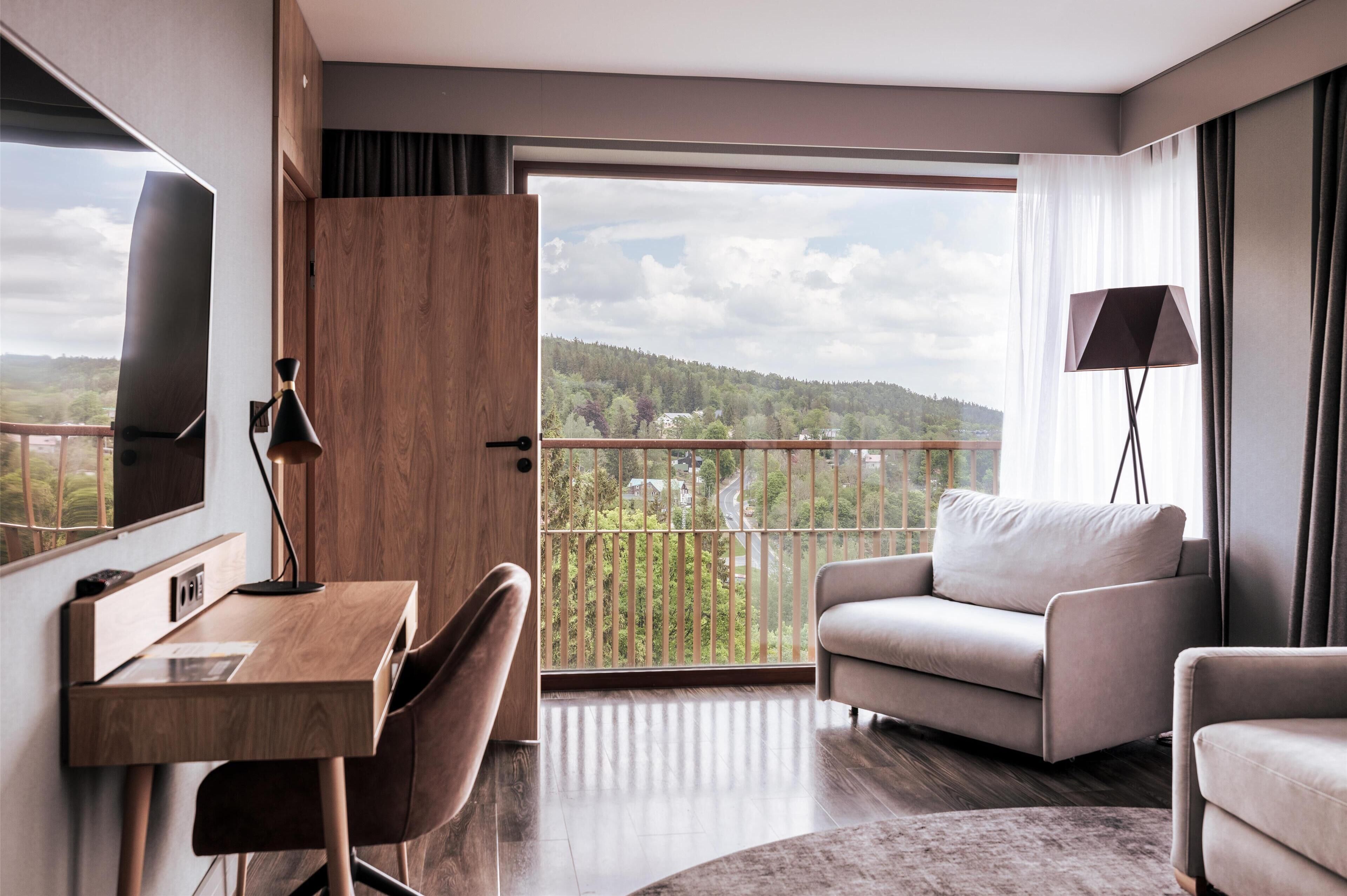 A modern hotel room with a wooden desk, a comfortable sofa, a TV, and a balcony with a scenic view.