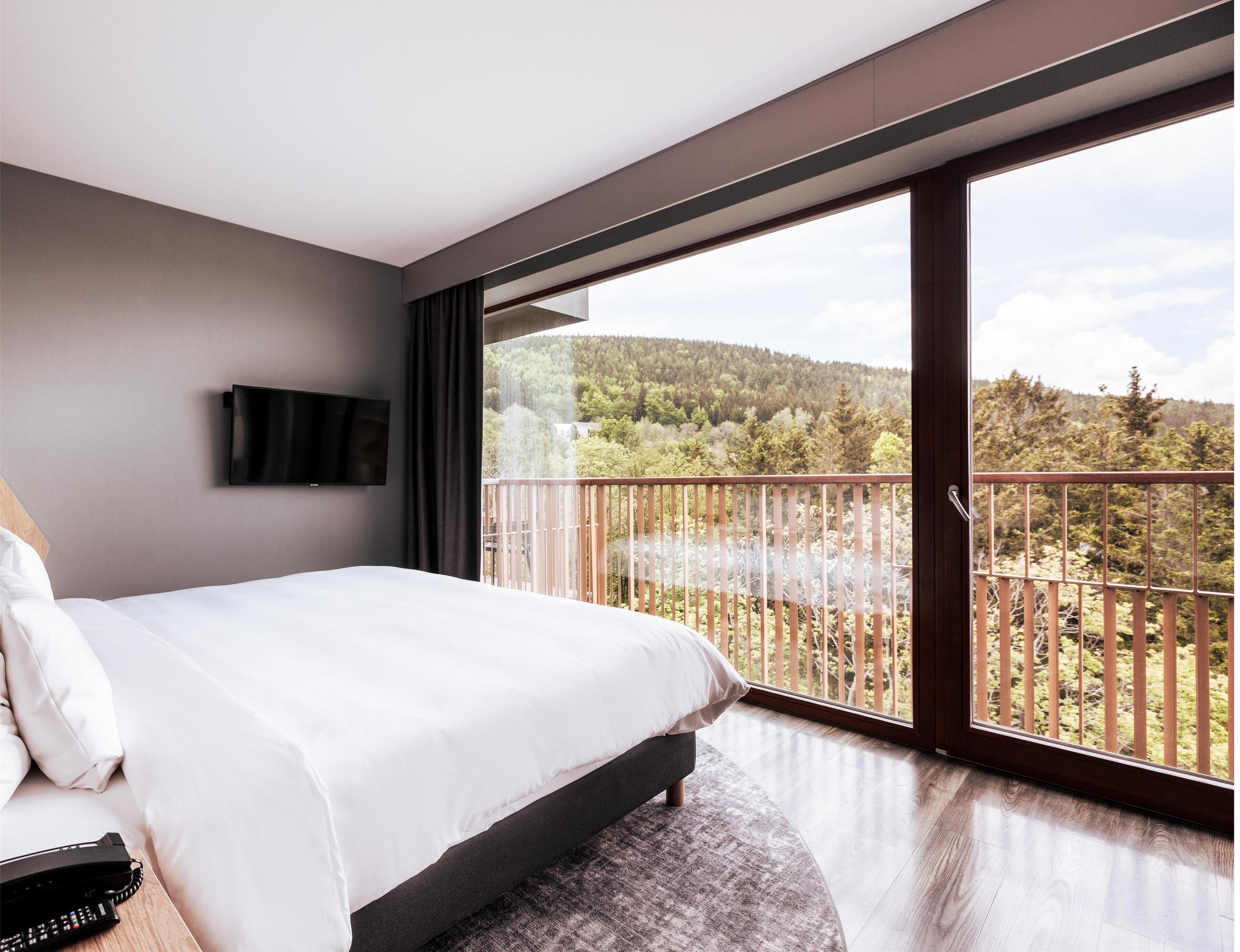 A modern bedroom with a large bed, a flat-screen TV mounted on the wall, and a balcony with a view of a forested hillside.