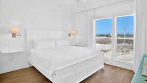 Ein helles Schlafzimmer mit einem großen Bett, zwei Nachttischlampen, einem Deckenventilator und Blick auf den Strand durch große Fenster.