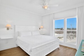 Un dormitorio blanco con una cama grande, dos lámparas de noche, un ventilador de techo y vista a la playa a través de amplias ventanas.