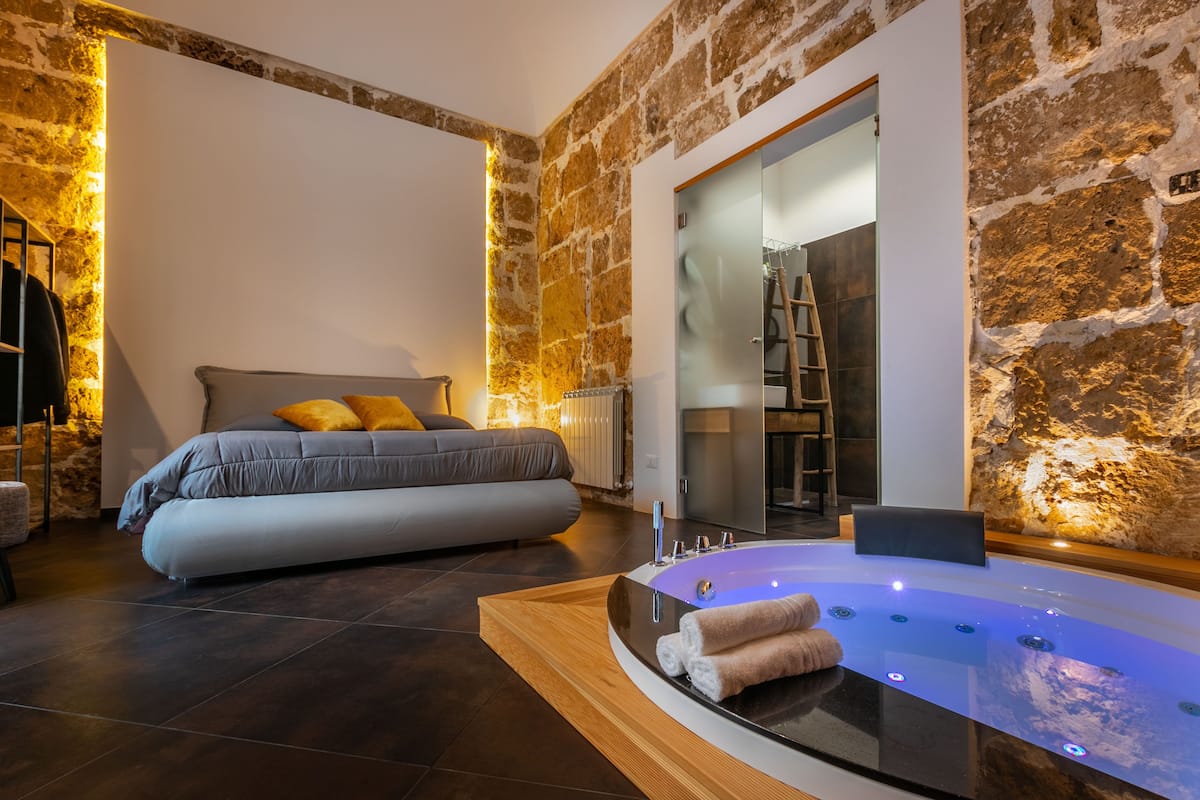 A modern bedroom with a large bed, a glass-enclosed bathroom with a bathtub, and stone walls.