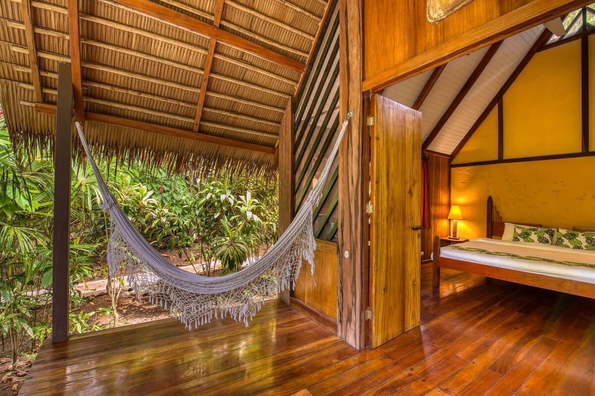 Un intérieur en bois avec un hamac, un lit et une vue sur la verdure.