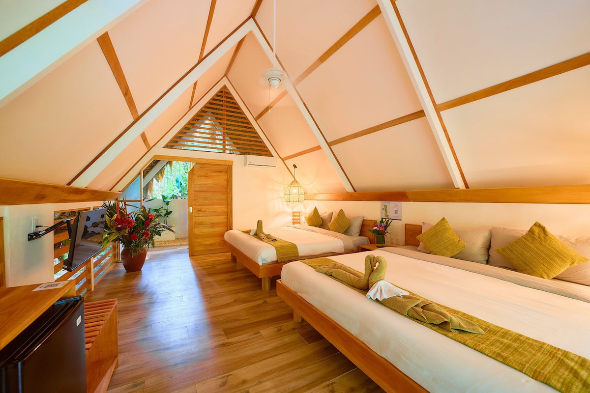 Une chambre mansardée chaleureuse et bien éclairée, avec des poutres en bois, deux lits recouverts de serviettes vertes, un bureau en bois et une plante en pot.