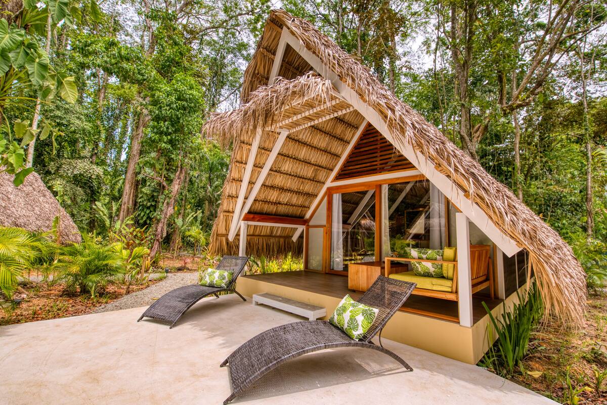 Un bungalow au toit de chaume, doté d’une porte en verre, de deux fauteuils de jardin en osier et d’une petite table en bois.