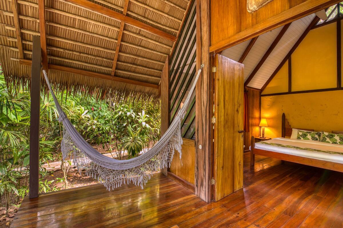 Un hamac sous un toit de chaume, un lit en bois avec vue sur une végétation luxuriante et un plancher en bois.