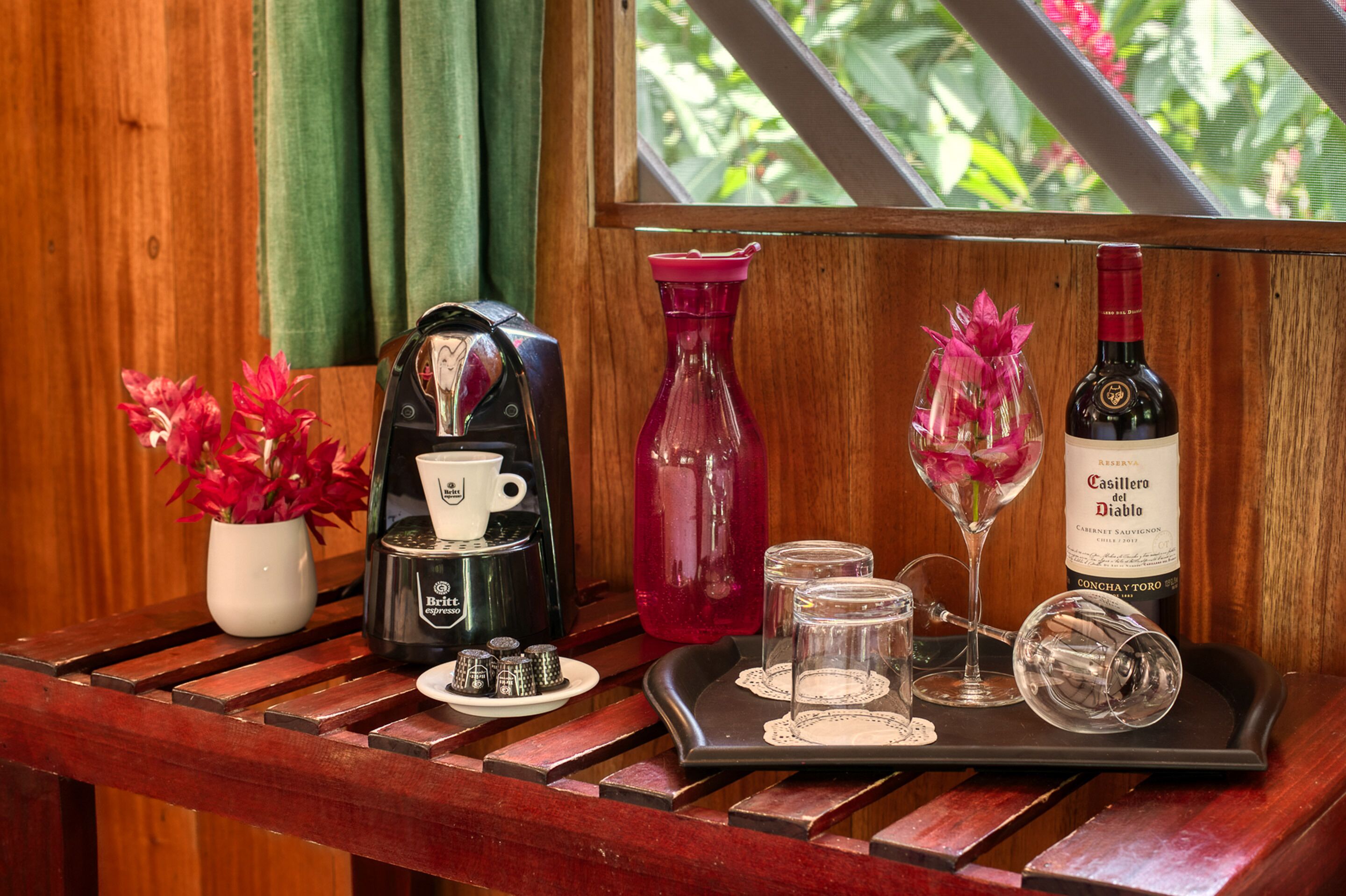 A tray with a coffee machine, a red vase, a wine bottle, and glasses on a wooden table.