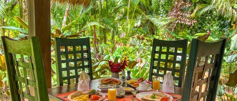 Una mesa de madera preparada para el desayuno, con platos de comida, un florero y un paisaje tropical.