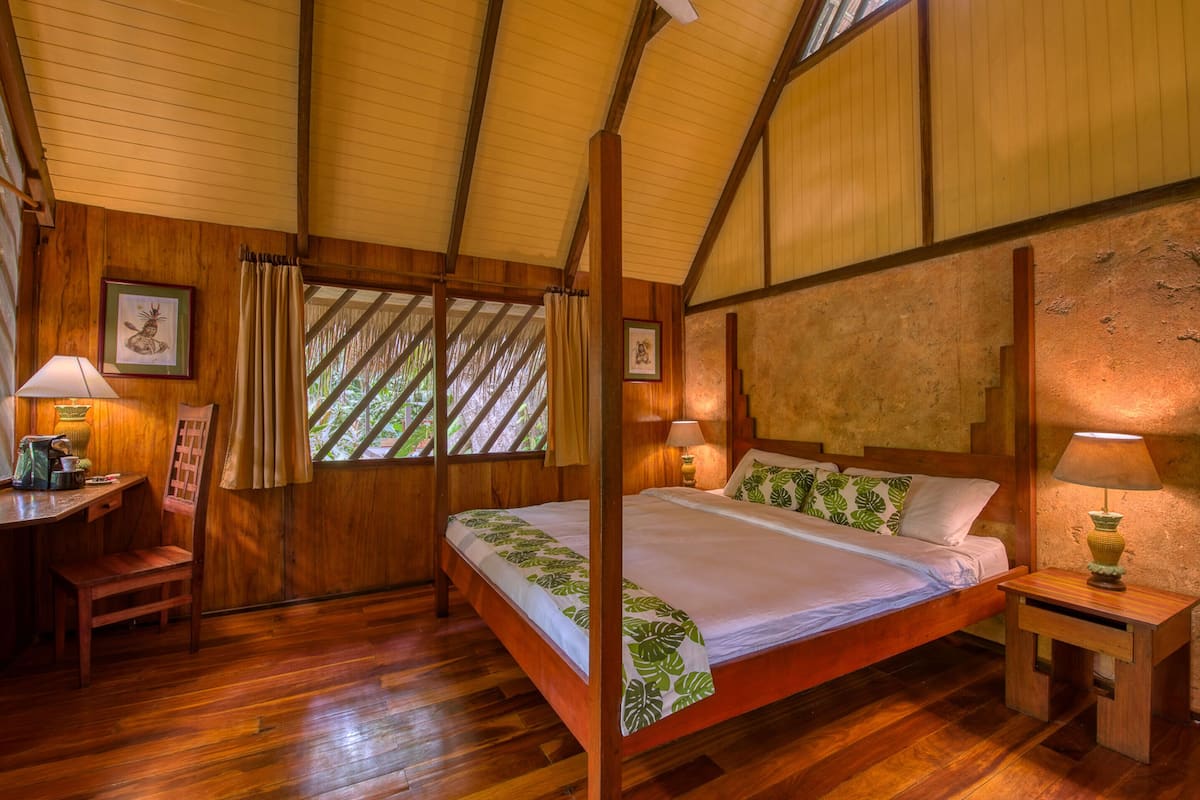 Un lit à baldaquin en bois, surmonté d’une couvre-lit à motifs de feuilles vertes, deux lampes de chevet et un bureau en bois avec une chaise.