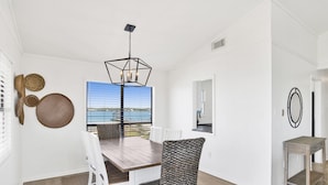 En matplats med ett träbord, vita stolar, en flätad stol och utsikt över havet genom stora fönster.