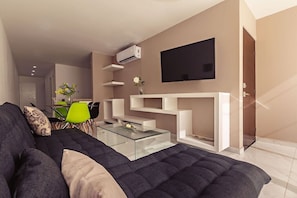 Ein modernes Wohnzimmer mit einem dunklen Sofa, einem Glastisch, einem Wand-Fernseher und einer integrierten, weißen Regaleinheit.