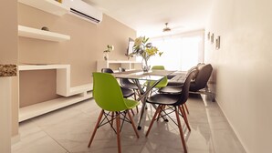 Uma área de refeições moderna com mesa de vidro, cadeiras verdes e pretas, e um vaso com flores.