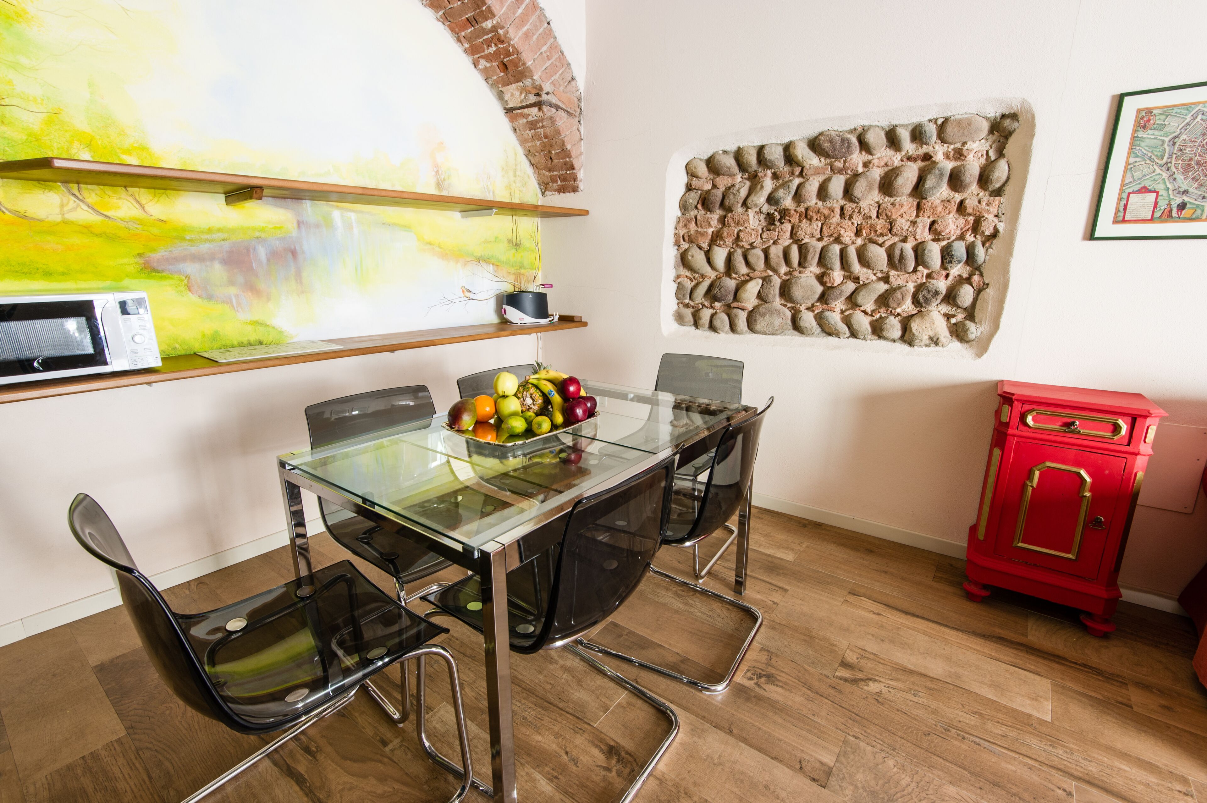 Un área de comedor con una mesa de vidrio, sillas negras, un frutero, un microondas, un armario rojo y una obra de arte en la pared de piedra.