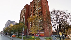 A tall red brick apartment building with multiple balconies, a sidewalk, and autumn trees. - Rupa pod oblacima (Zagreb)