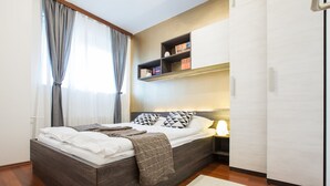 A modern bedroom with a wooden bed, a bedside table with a lamp, a bookshelf, and a large window with curtains. - Rupa pod oblacima (Zagreb)