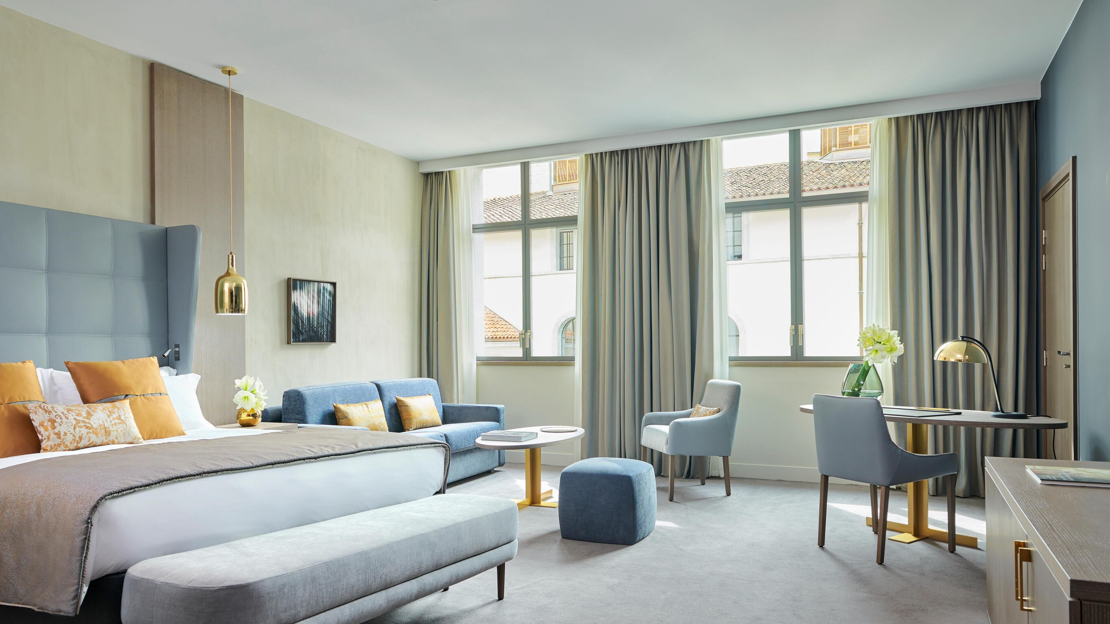 A modern hotel room with a large bed, a sofa, a desk, and a chair. There are curtains on the windows, and a painting on the wall.