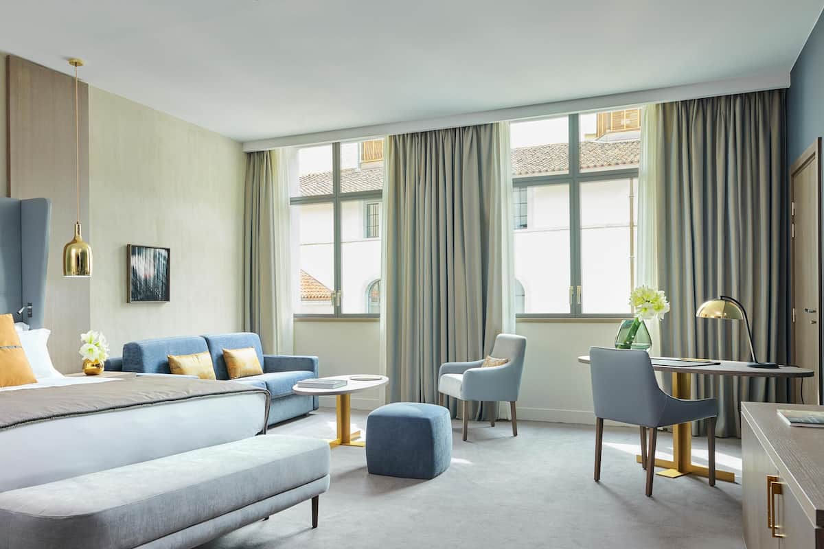A modern hotel room with a large bed, a sofa, a desk, and a chair. There are curtains on the windows, and a painting on the wall.