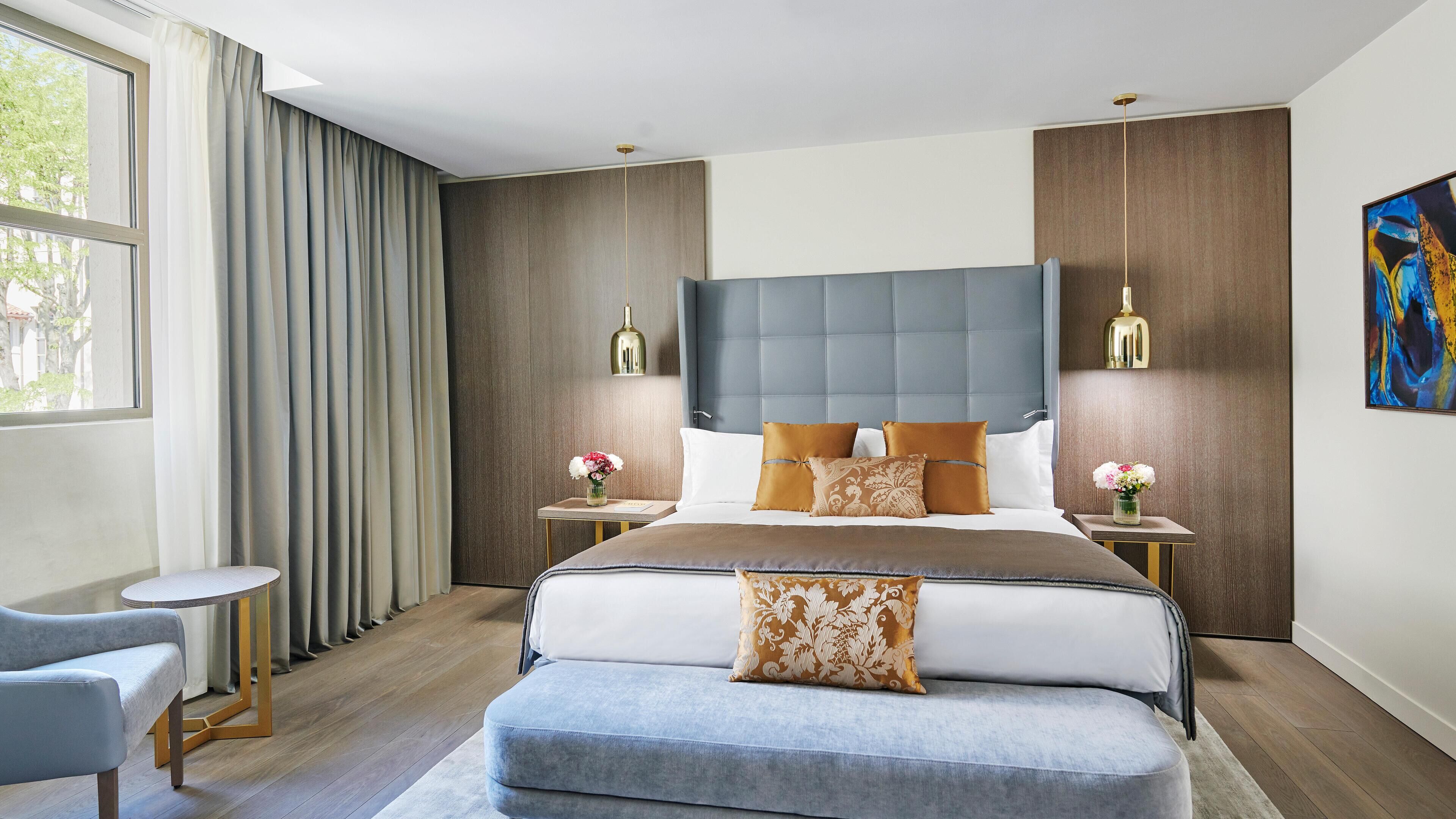 A modern hotel bedroom with a large bed, two bedside tables, a small round table, a sofa, and a large window with curtains.