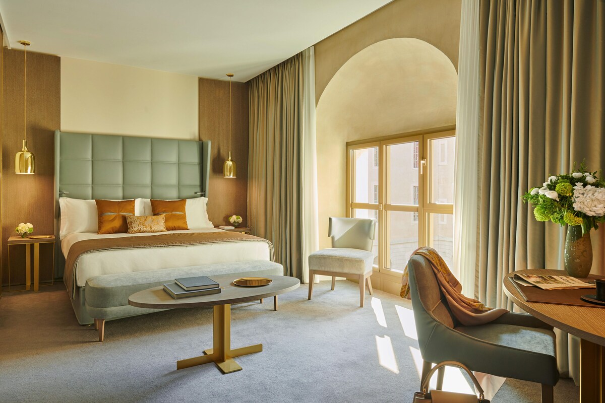 Habitación de hotel con una cama grande, una zona de estar con mesa y sillas, un escritorio con un jarrón de flores y una ventana con cortinas.