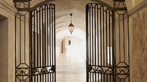 Una imponente entrada abovedada con puertas metálicas decorativas que dan a un pasillo con piso de piedra y un candelabro colgante.
