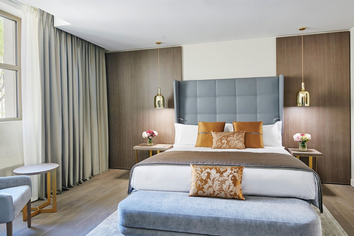 Un dormitorio moderno con una cama grande, dos mesitas de noche, una mesita redonda pequeña y un banco.