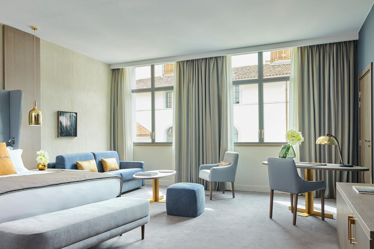Una habitación de hotel moderna con una cama grande, un sofá, un escritorio y una silla. Hay cortinas en las ventanas y un cuadro en la pared.