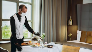 Habitación de hotel con una mesa de comedor dispuesta para una comida, una cama con almohada marrón y una ventana con cortinas.
