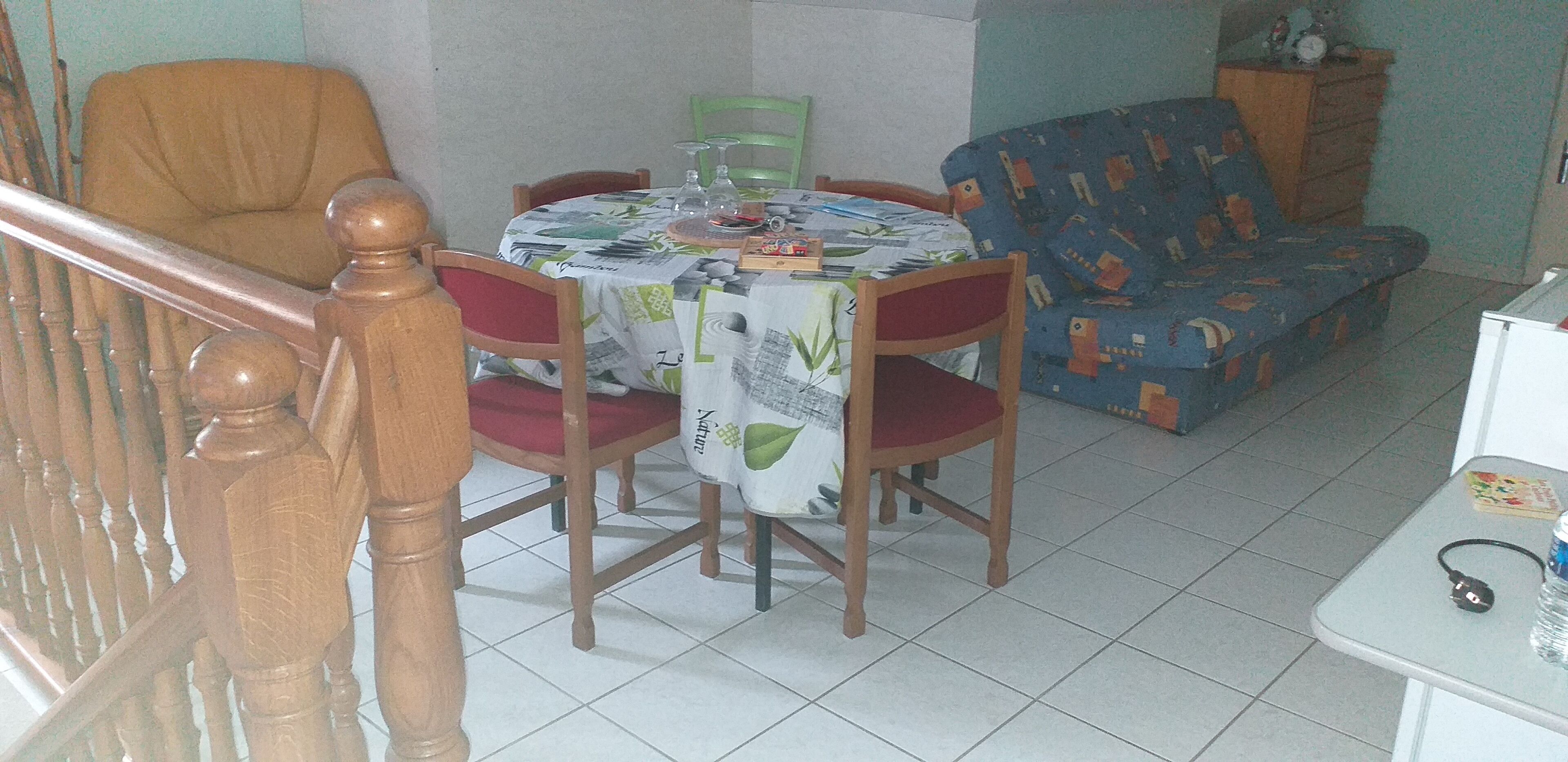 A dining area with a table set for four, surrounded by wooden chairs with red cushions. A patterned sofa is against the wall, and a small table with a bottle and a power cord is visible.